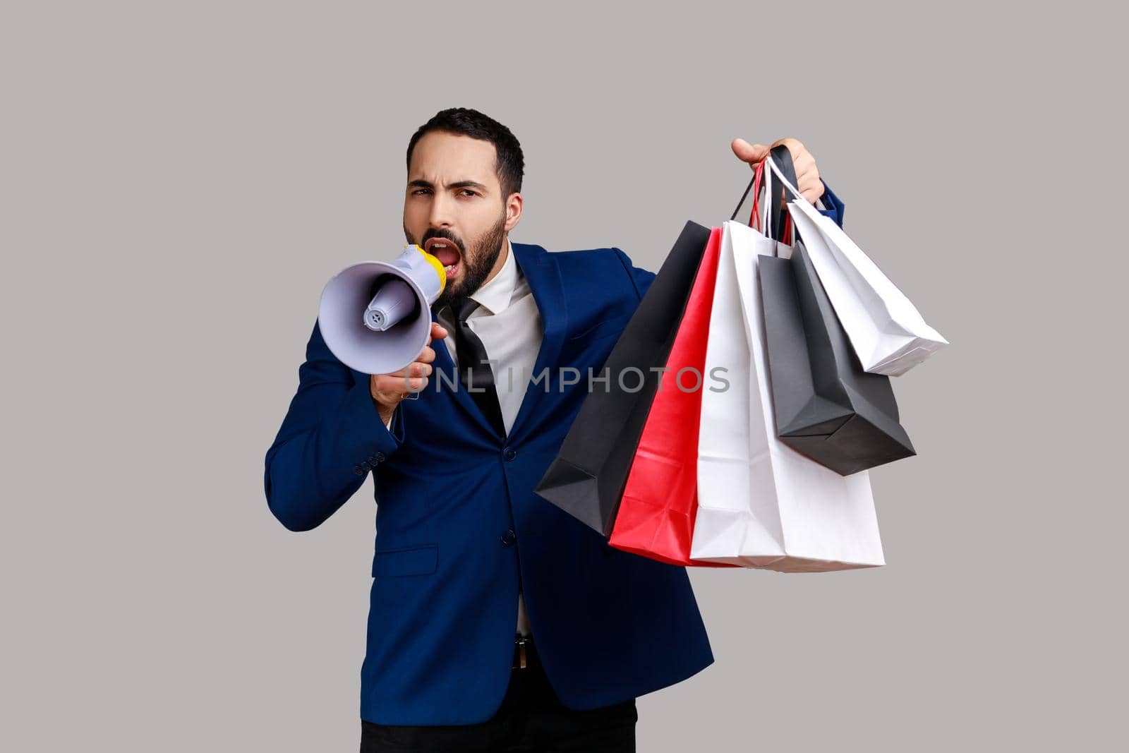 Bearded man announcing shopping discounts and sale, holding shopping bags with purchases, screams in loud speaker, wearing official style suit. Indoor studio shot isolated on gray background.