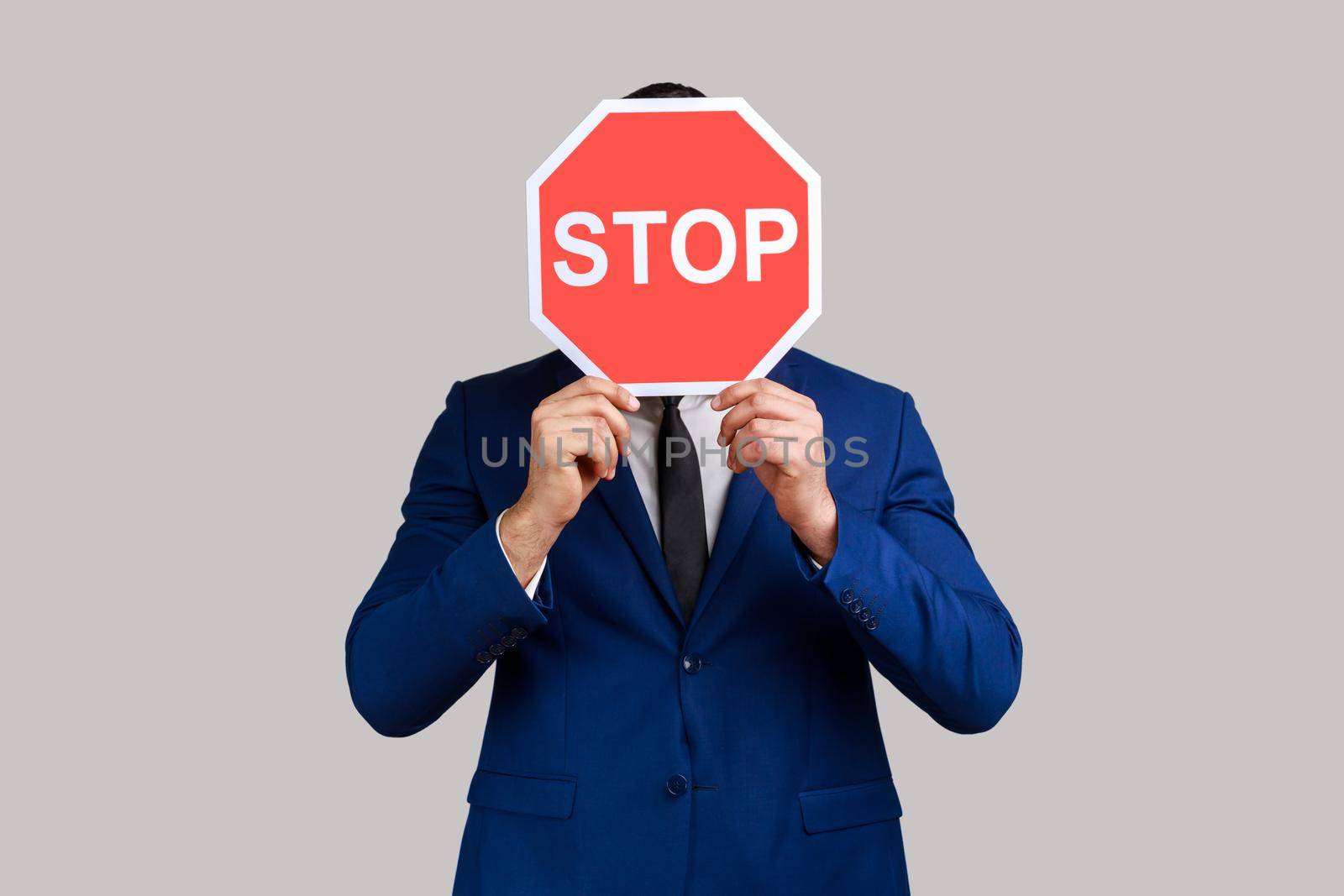 Portrait of man hiding face behind Stop symbol, red traffic sign warning of restricted access, banned service, wearing official style suit. Indoor studio shot isolated on gray background.