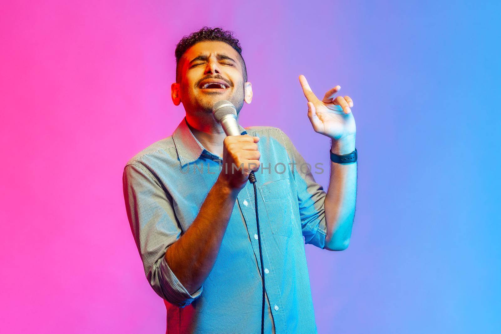 Portrait of happy man in shirt loudly singing song holding microphone in hand, having fun resting in karaoke, singer performance. Indoor studio shot isolated on colorful neon light background.