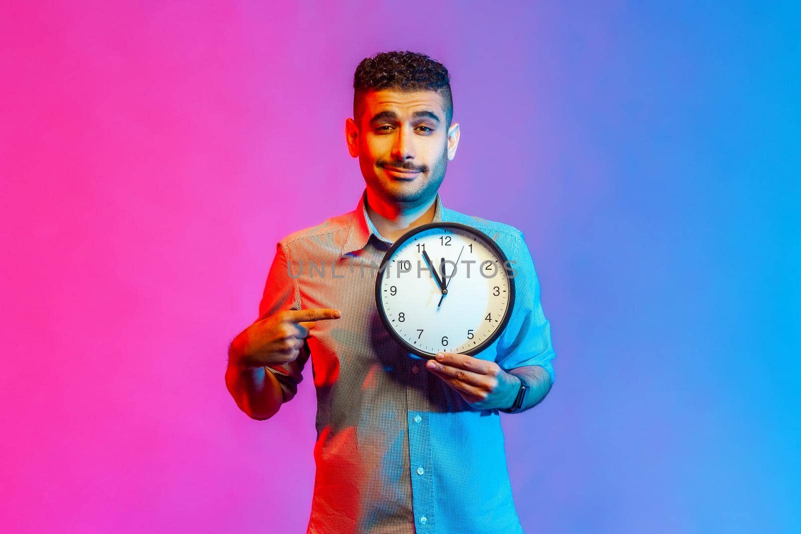 Look at time. Portrait of handsome positive man in shirt pointing big clock and looking at camera, showing wall watches to hurry up. Indoor studio shot isolated on colorful neon light background.