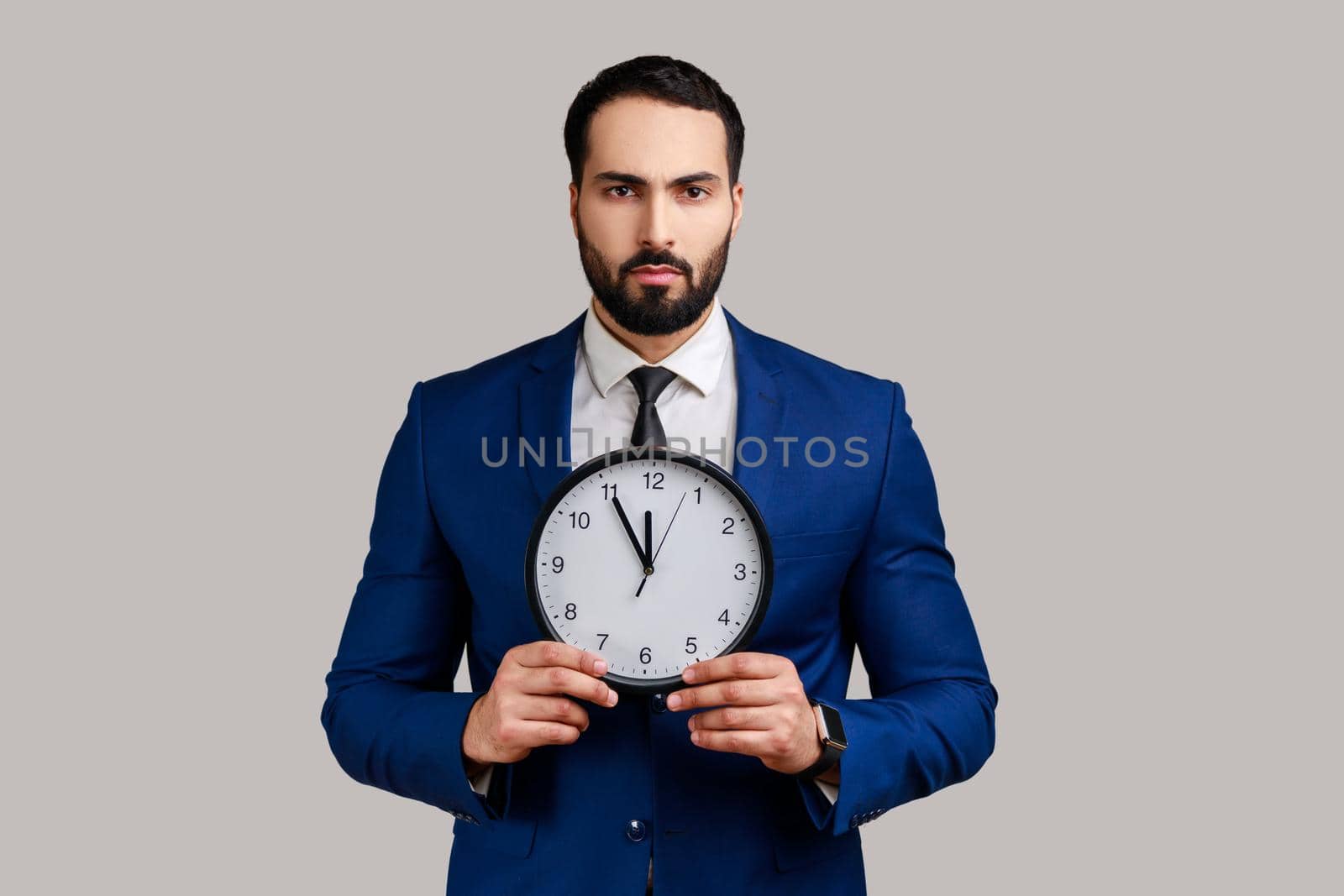 Portrait of serious bossy bearded man afraid of being late, holding in hand wall watch, deadline, punctuality, wearing official style suit. Indoor studio shot isolated on gray background.