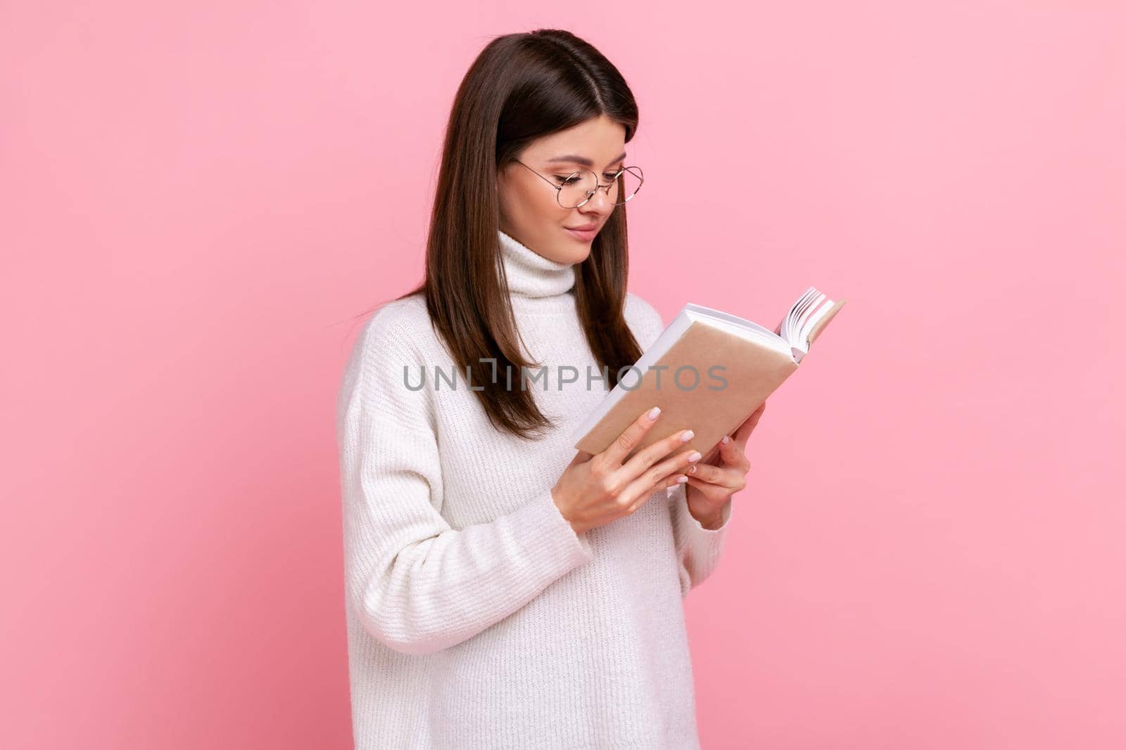 Concentrated smiling brunette girl holds book, enjoys reading, standing absorbed with exciting plot, wearing white casual style sweater. Indoor studio shot isolated on pink background.