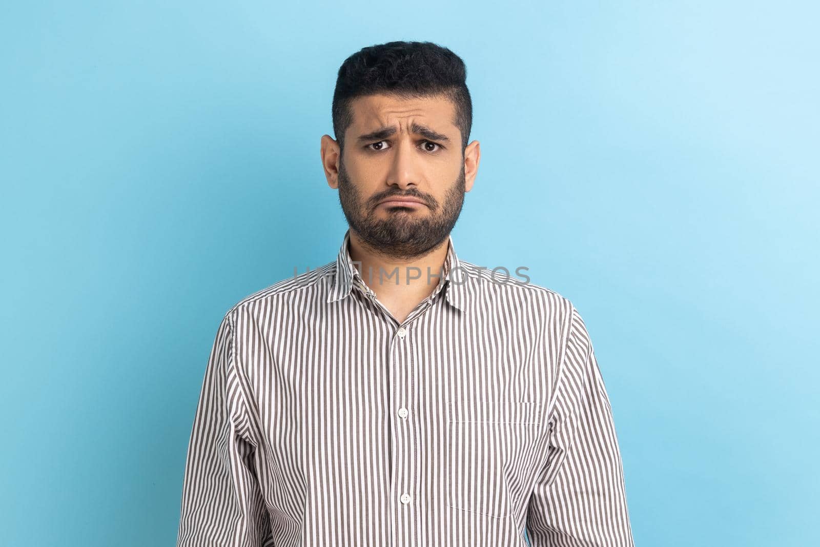 Bearded upset businessman standing and looking at camera with dissatisfied sadness face, expressing sorrow, having bad mood, wearing striped shirt. Indoor studio shot isolated on blue background.