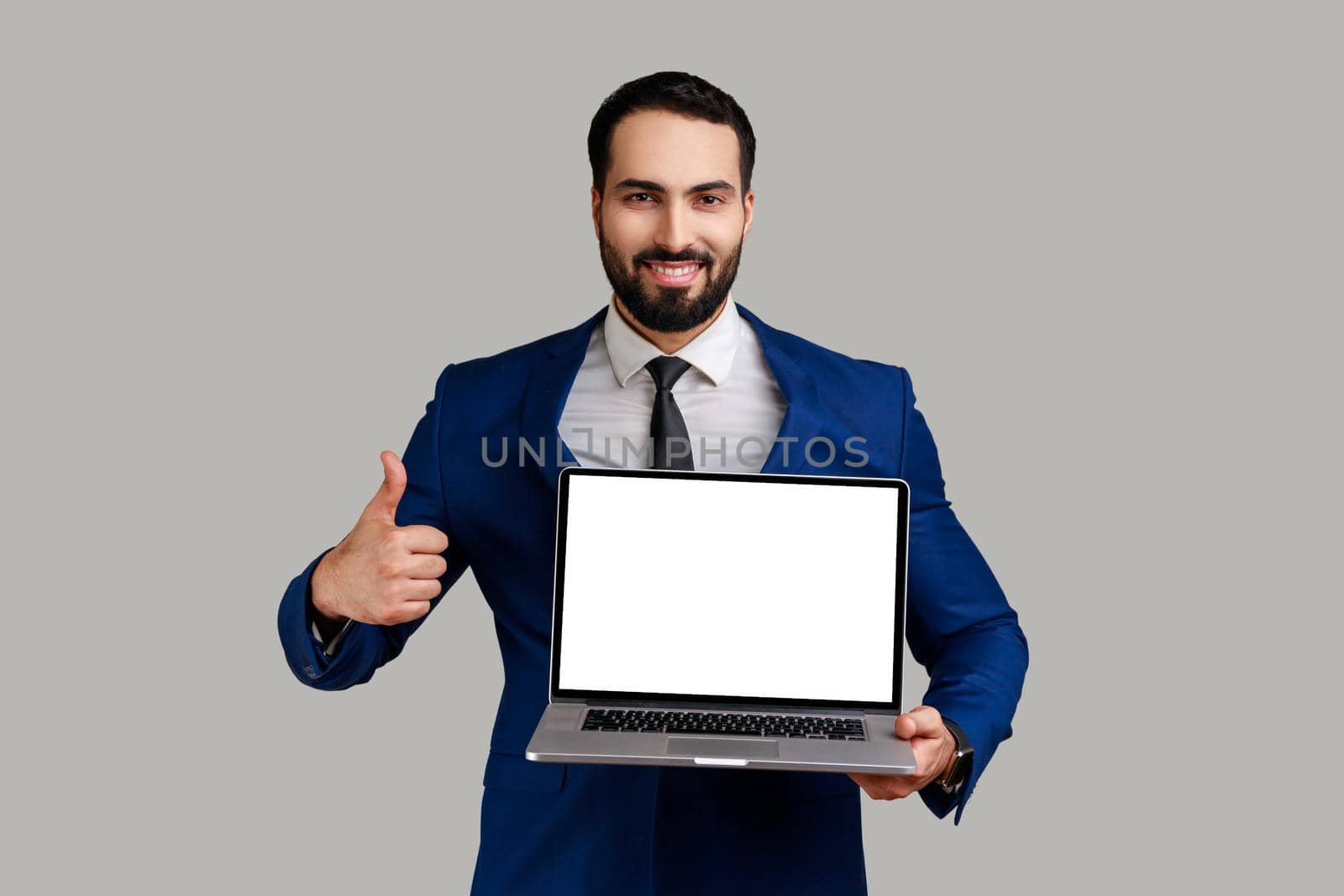 Portrait of satisfied delighted bearded man working on laptop with smile and showing thumb up, looking at camera, wearing official style suit. Indoor studio shot isolated on gray background.