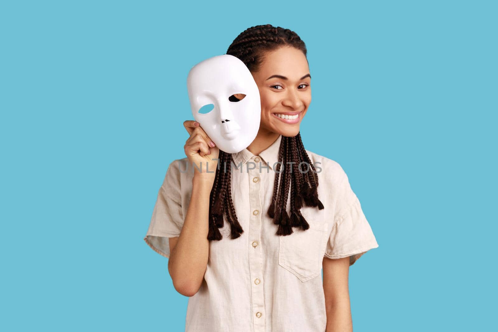 Woman with dreadlocks removing white mask from face showing his smiling expression, good mood, pretending to be another person, wearing white shirt. Indoor studio shot isolated on blue background.