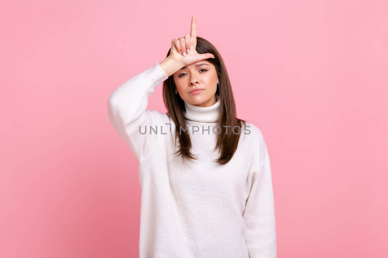 Depressed woman showing loser gesture, L finger sign on forehead, upset about dismissal, unlucky day, wearing white casual style sweater. Indoor studio shot isolated on pink background.