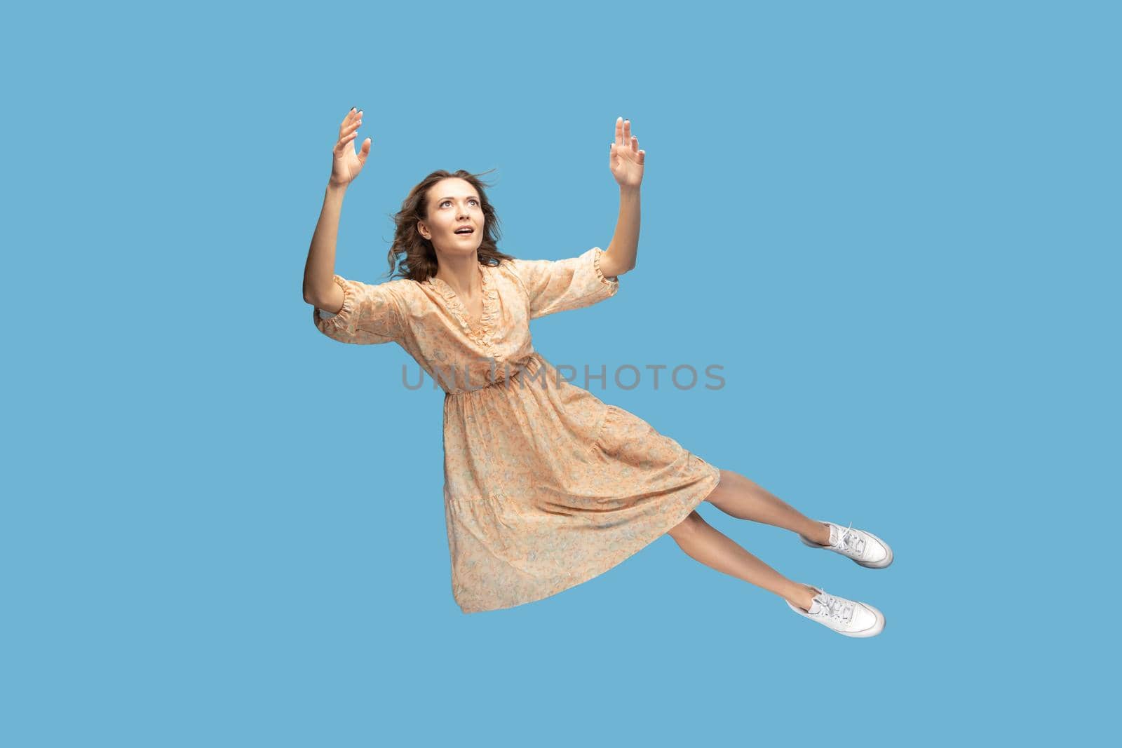 Hovering in air. Full length beautiful pensive girl in yellow dress levitating flying in mid-air, looking up with dreamy relaxed expression. indoor studio shot isolated on blue background