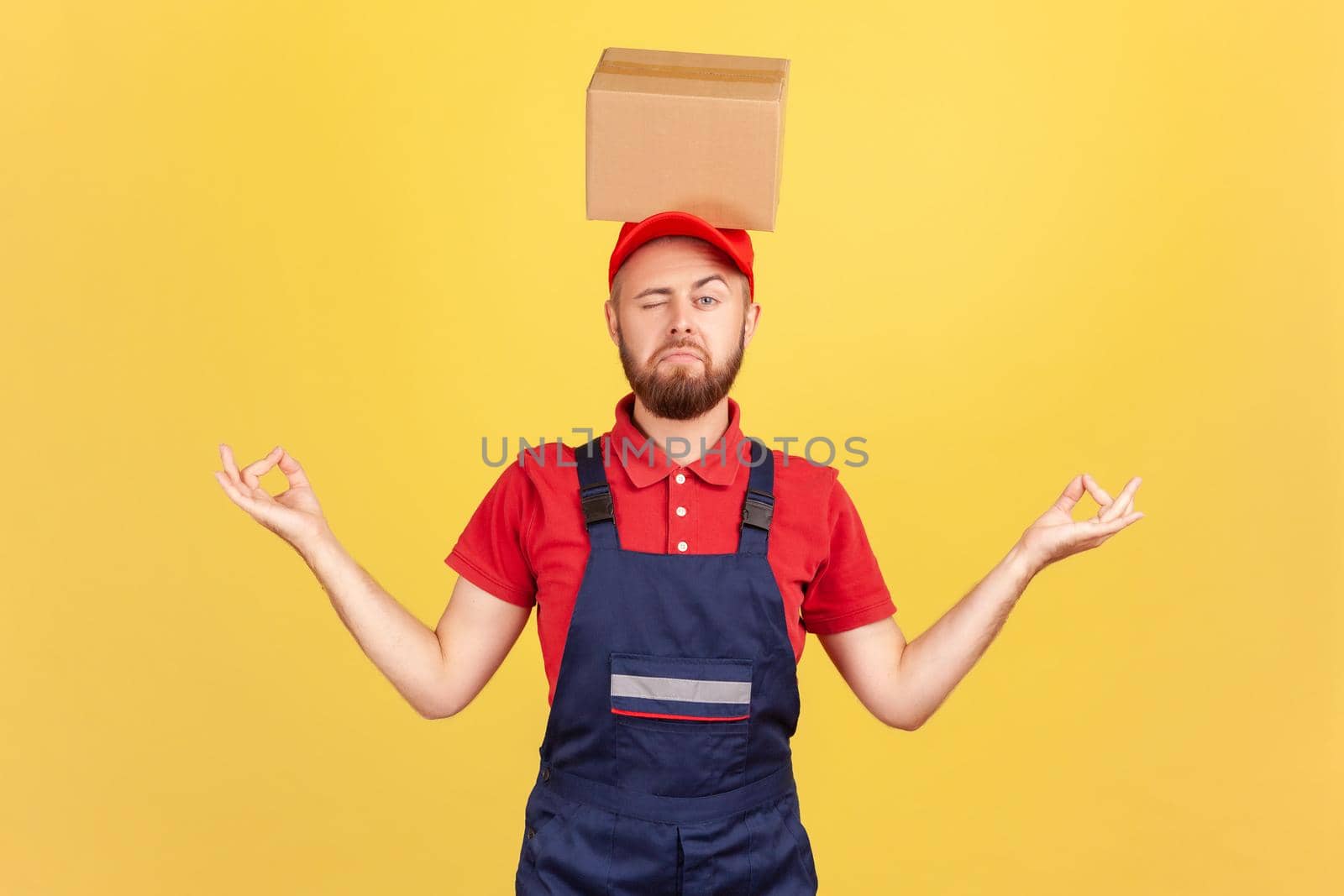 Portrait of relaxed courier man standing with cardboard box on his head, practicing yoga, looking at camera with one eye, calms down. Indoor studio shot isolated on yellow background.
