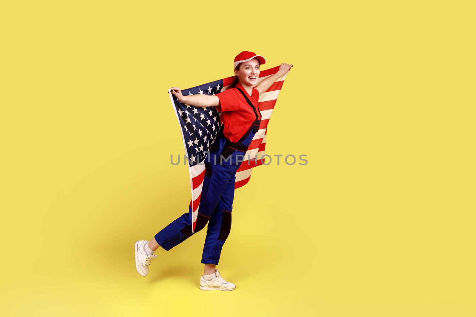 Full length portrait of happy positive worker woman posing with american flag in hands, celebrating patriotic holiday, wearing overalls and red cap. Indoor studio shot isolated on yellow background.