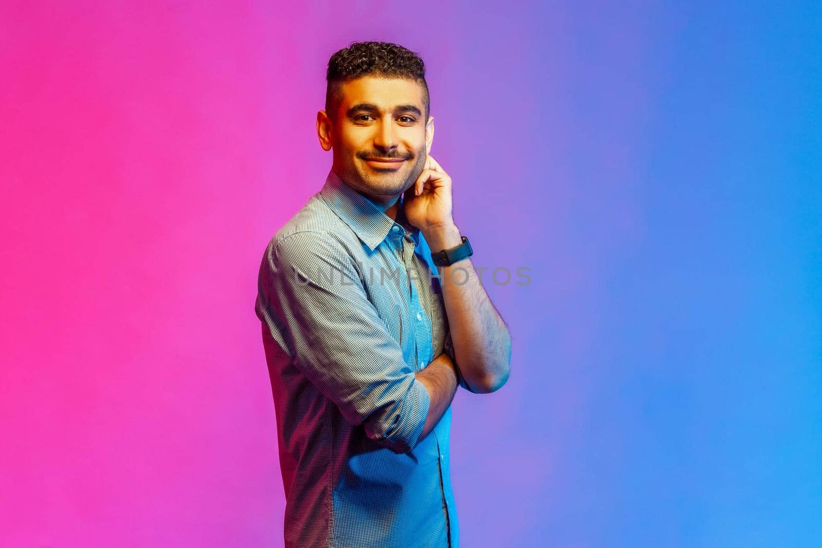 Portrait of young adult smiling handsome man in shirt standing looking at camera with happy positive facial expression. Indoor studio shot isolated on colorful neon light background.