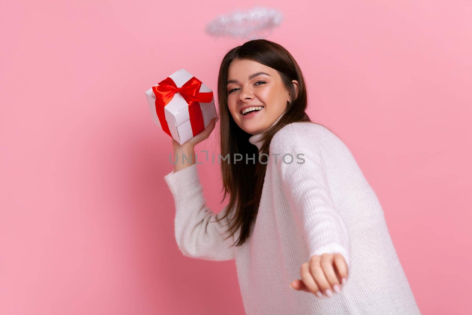 Satisfied angelic woman with nimb over her head, throwing present box, congratulating with holiday, wearing white casual style sweater. Indoor studio shot isolated on pink background.