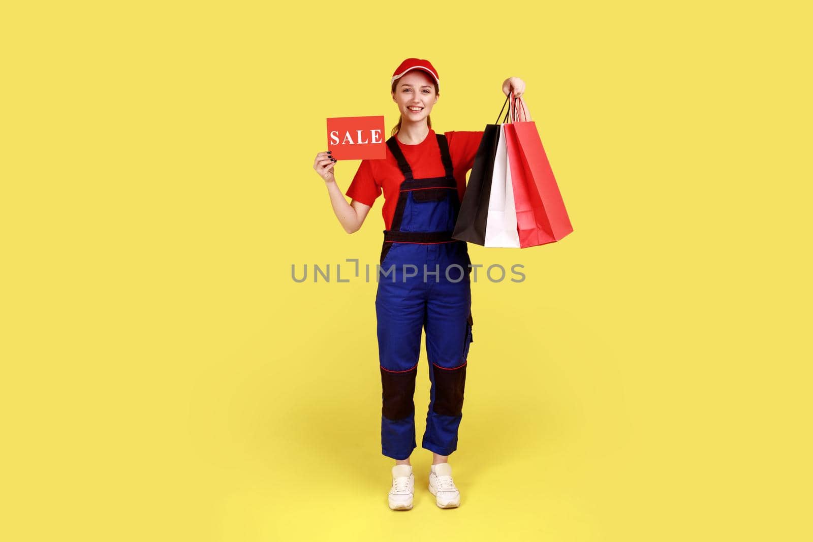 Full length portrait of delivery woman standing with shopping bags in hands, and showing card with sale inscription, wearing overalls and red cap. Indoor studio shot isolated on yellow background.