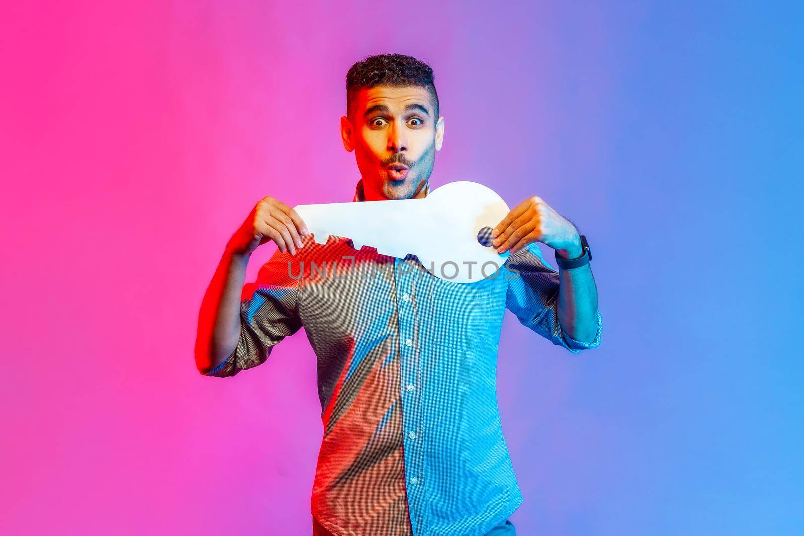Portrait of astonished man in shirt holding huge paper key and being amazed to get new home, real estate purchase, rental service. Indoor studio shot isolated on colorful neon light background.