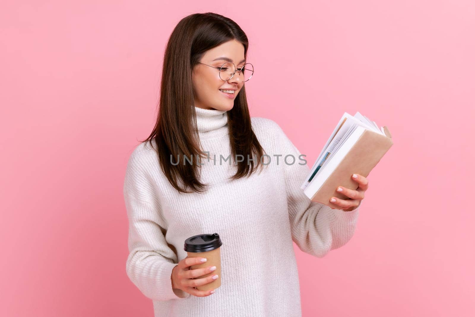 Side view of satisfied female in glasses reading interesting book and enjoying hot take away coffee, wearing white casual style sweater. Indoor studio shot isolated on pink background.