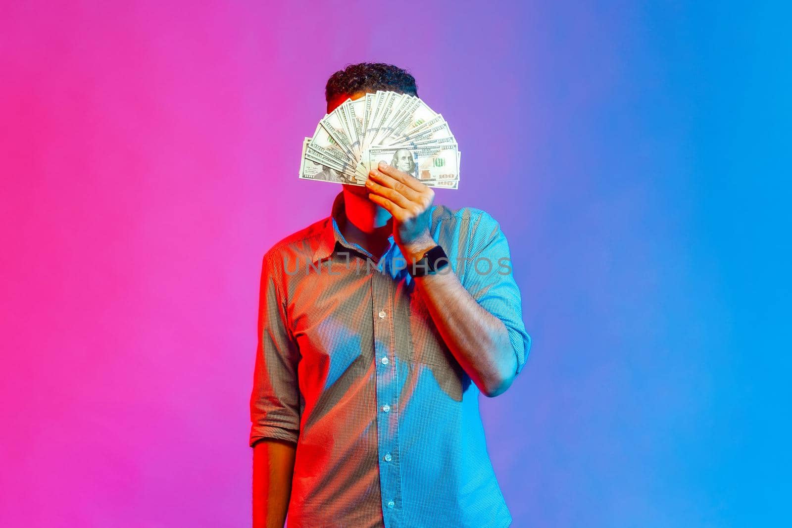 Portrait of man in shirt hiding face behind bunch of dollar banknotes, anonymous person holding money, lottery win, big profit. Indoor studio shot isolated on colorful neon light background.