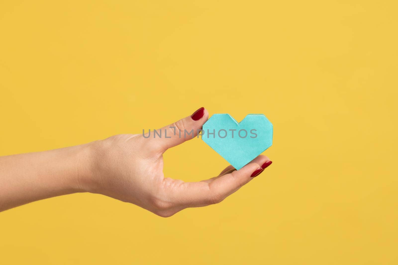 Profile side view closeup of woman hand holding tiny turquoise heart, symbol of love, showing romantic feelings. Indoor studio shot isolated on yellow background.