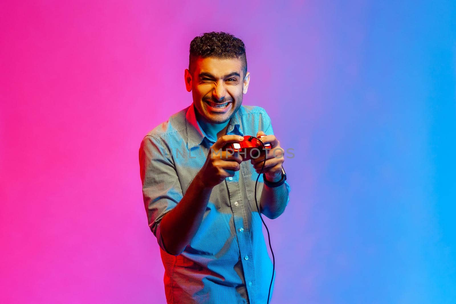 Portrait of amazed man in shirt holding in hands red gamepad joystick, looking at camera with optimistic expression, enjoying game. Indoor studio shot isolated on colorful neon light background.