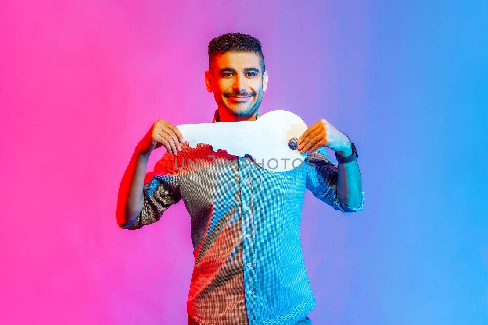 Portrait of positive man in shirt holding huge paper key and smiling satisfied with new home, real estate purchase, rental service. Indoor studio shot isolated on colorful neon light background.