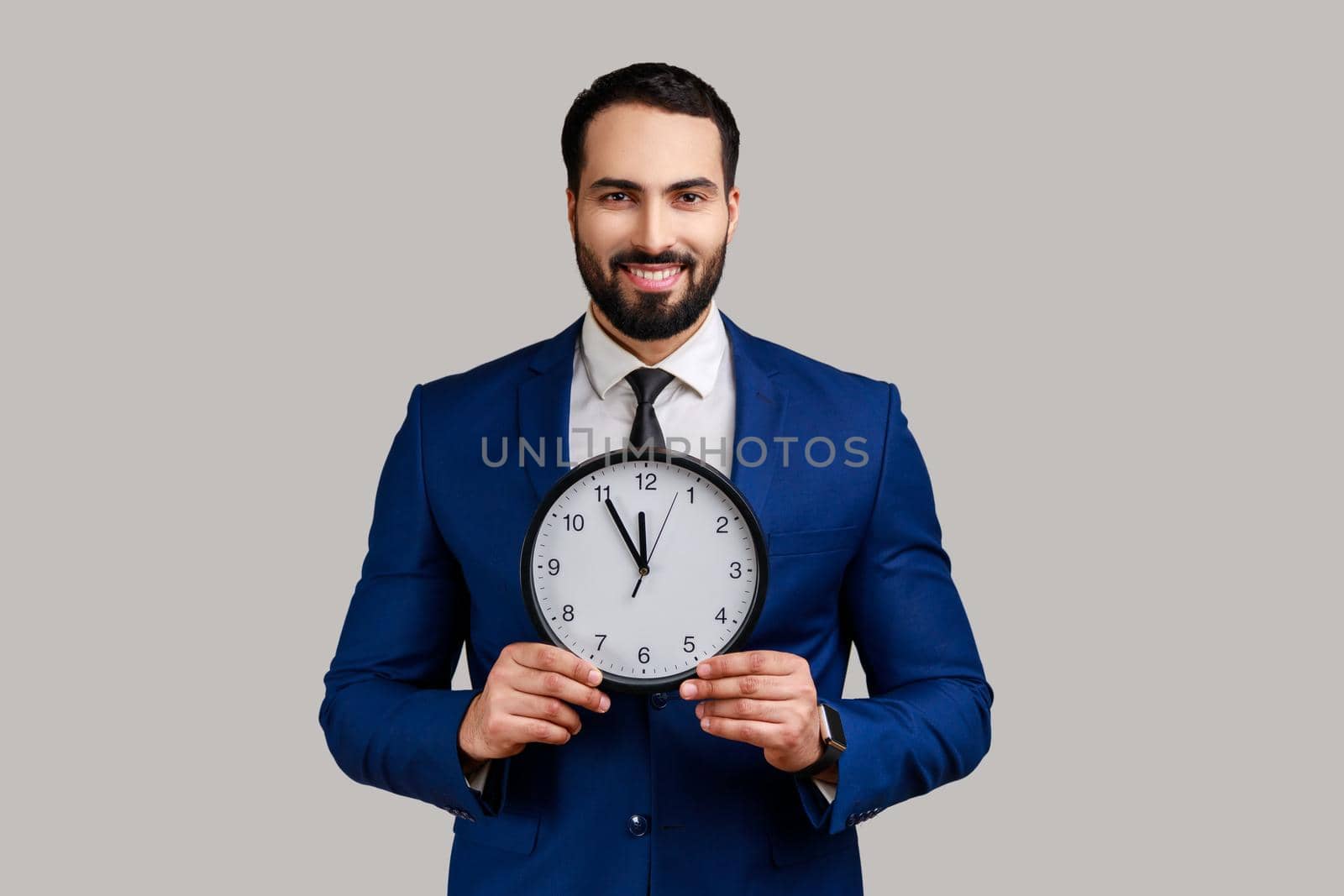 Handsome bearded businessman holding big clock and looking at camera with toothy smile, playful optimistic expression, wearing official style suit. Indoor studio shot isolated on gray background.