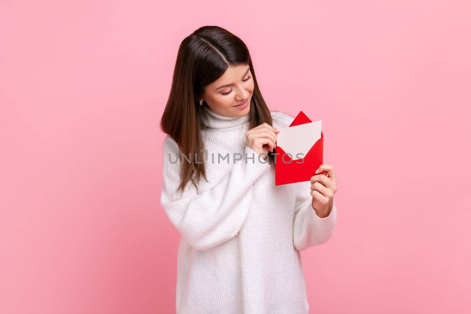 Portrait of happy woman holding red romantic envelope and looking at letter with smile and interest, wearing white casual style sweater. Indoor studio shot isolated on pink background.