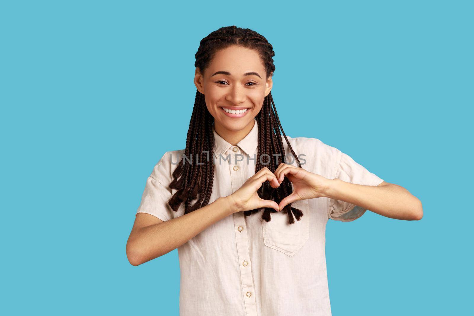 Beautiful woman with black dreadlocks gesture heart over chest, expresses love, says be my valenetine, smiles positively, wearing white shirt. Indoor studio shot isolated on blue background.