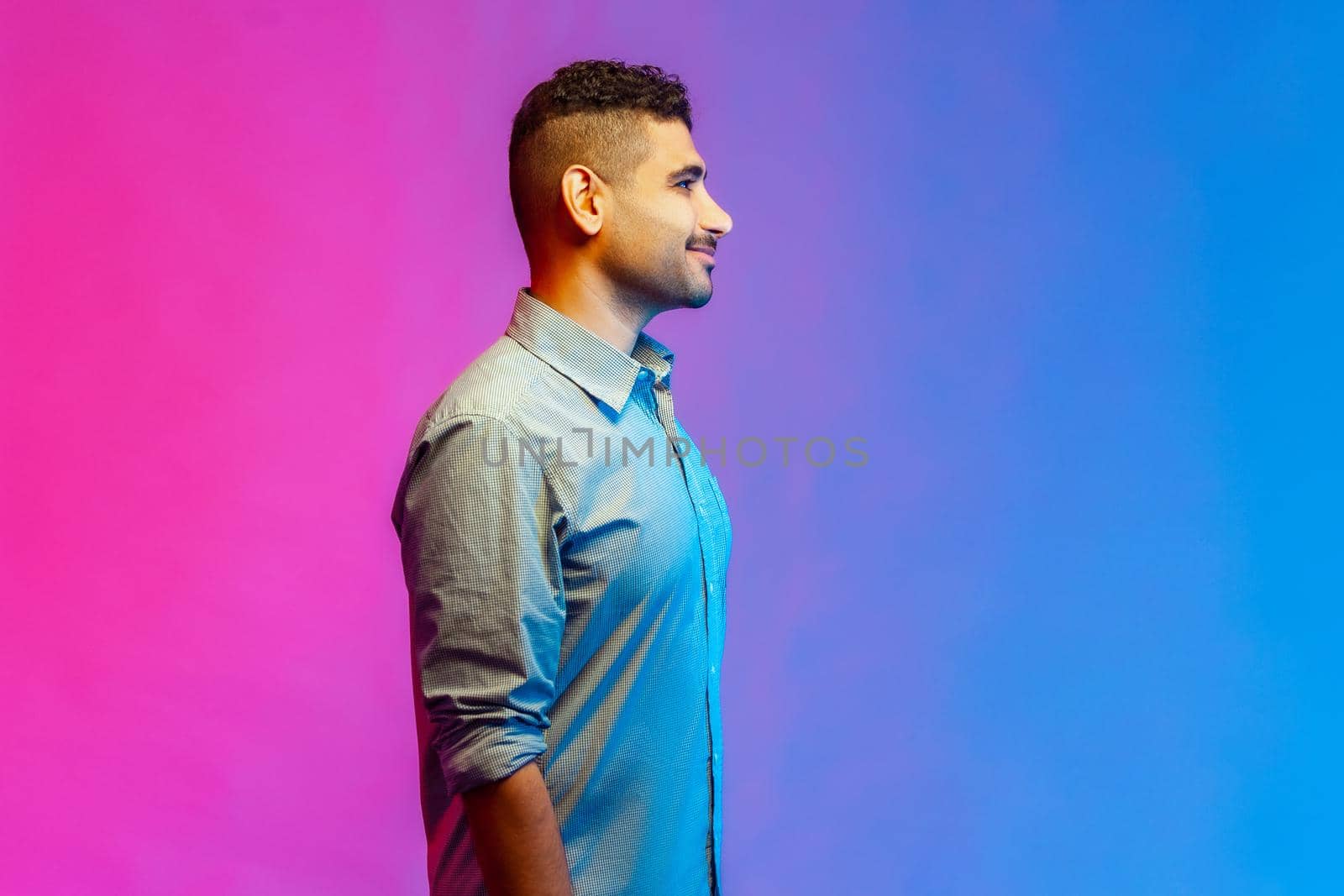 Side view portrait of young adult smiling handsome man in shirt standing looking ahead with happy positive facial expression. Indoor studio shot isolated on colorful neon light background.