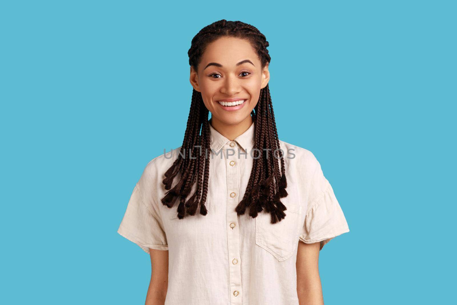 Portrait of cheerful beautiful woman with black dreadlocks, having charming engaging smile and positive emotions, wearing white shirt. Indoor studio shot isolated on blue background.