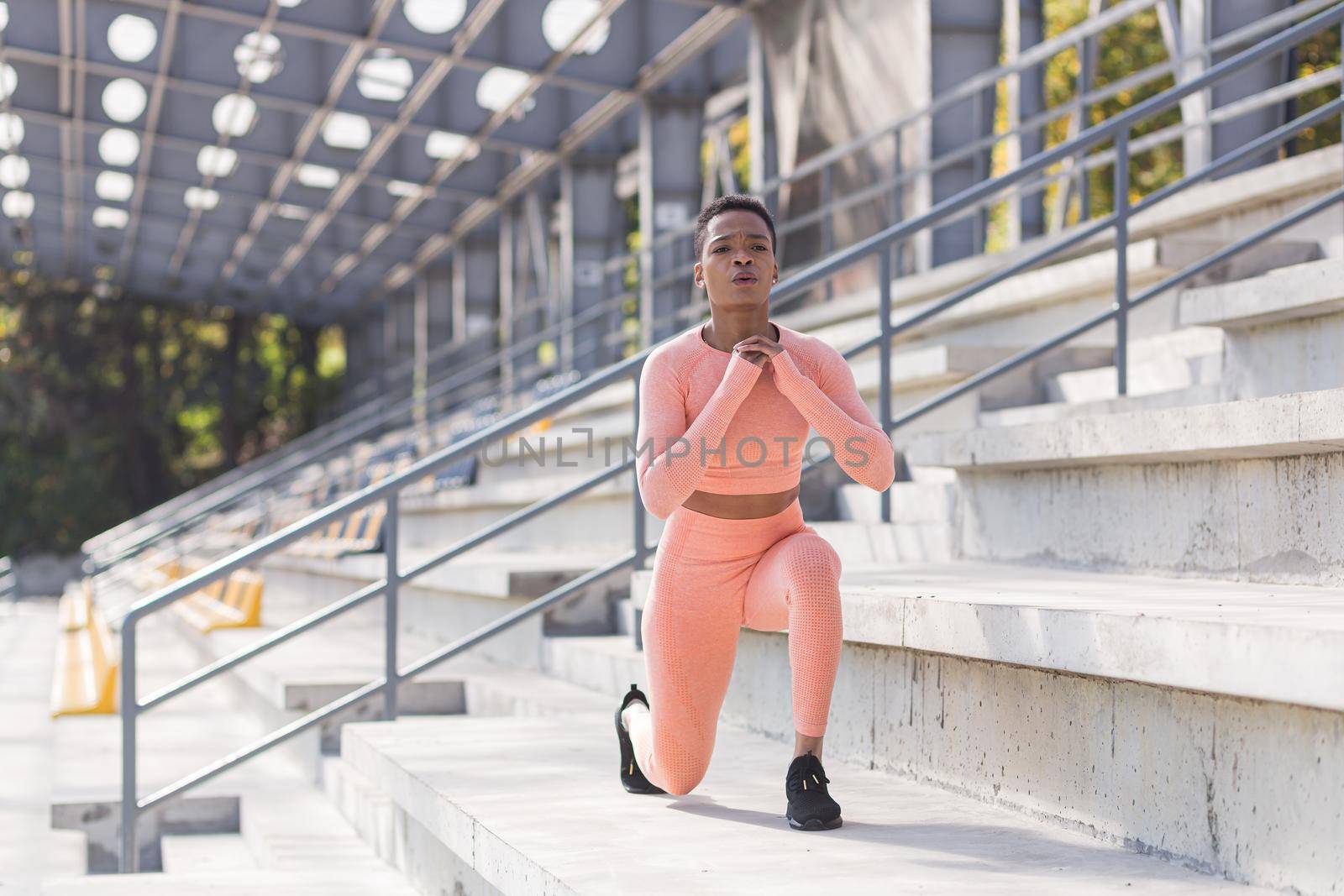 African American woman performs fitness exercises for weight loss, performs squats in a pink suit in the morning in the air near the stadium