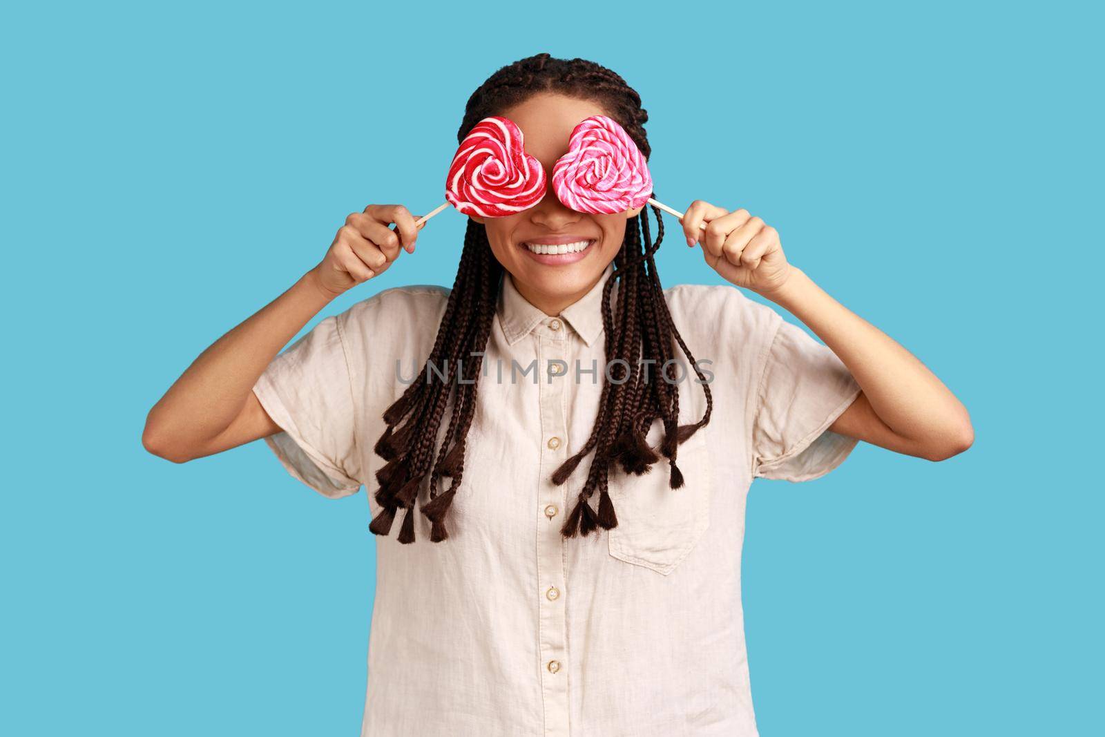 Portrait of playful childish romantic woman with black dreadlocks covering eyes with sugary hear shape candies, wearing white shirt. Indoor studio shot isolated on blue background.