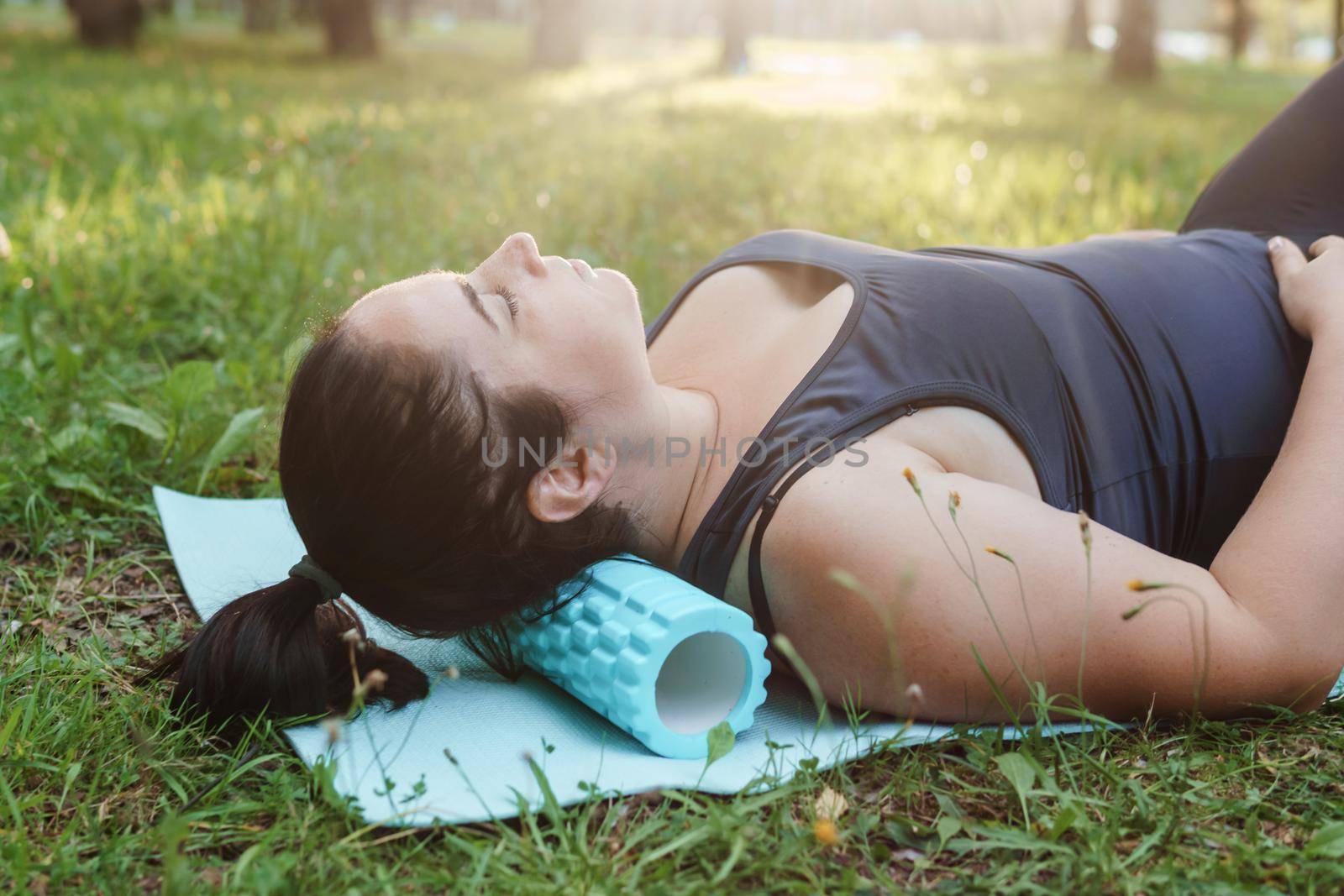 A charming brunette woman plus-size body positive practices sports in nature. Woman is engaged with a massage roller for the body in the park on a sports mat.