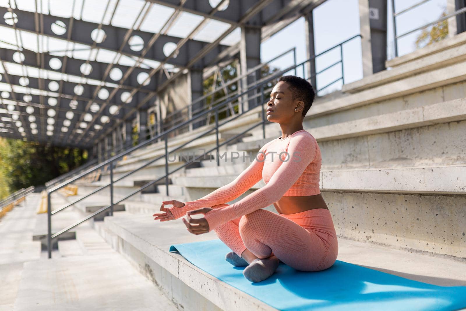 Young beautiful woman in a pink tracksuit meditating in the stadium in the morning, fitness trainer resting after training