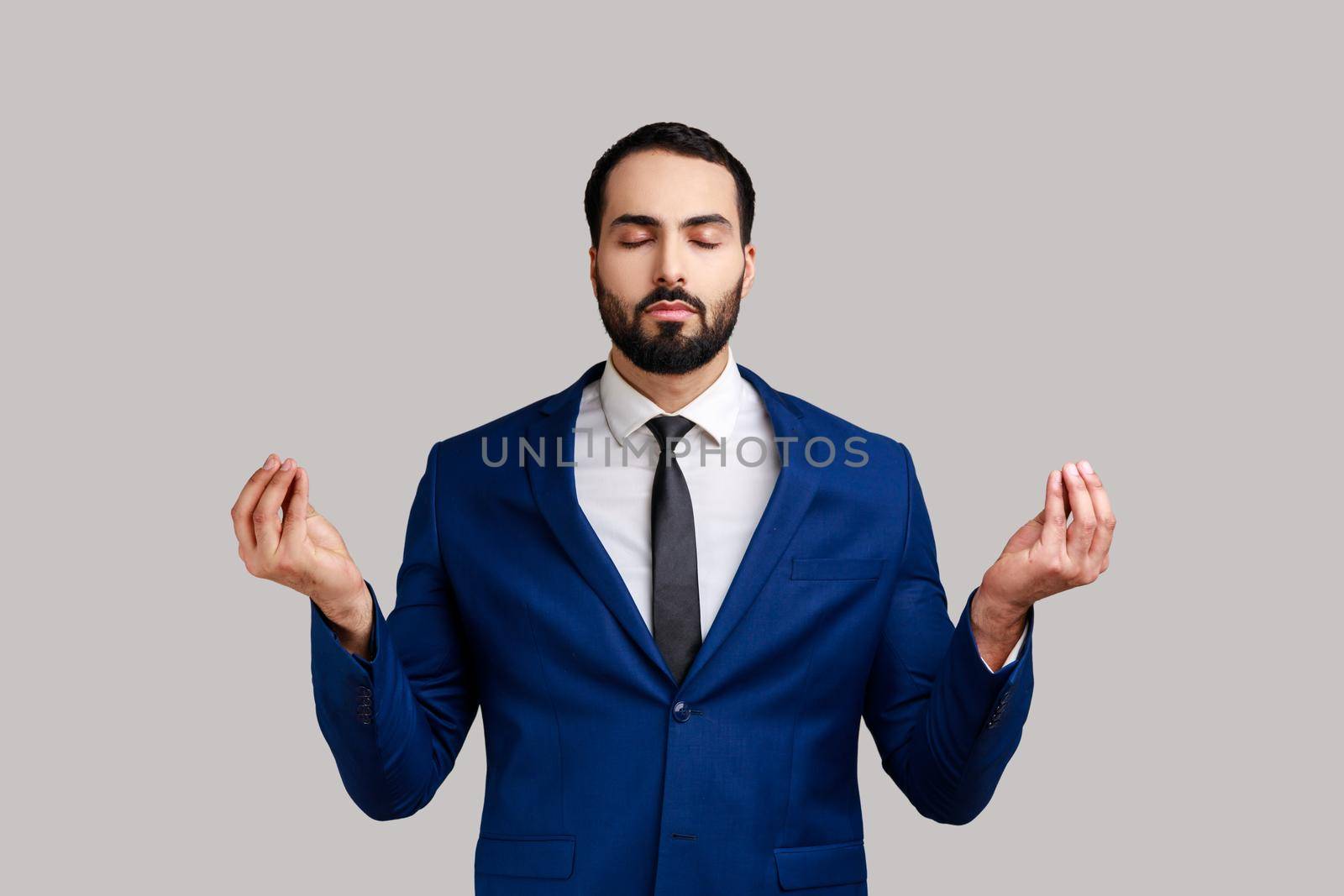 Portrait of relaxed handsome bearded man standing with raised arms and doing yoga meditating exercise, wearing official style suit. Indoor studio shot isolated on gray background.