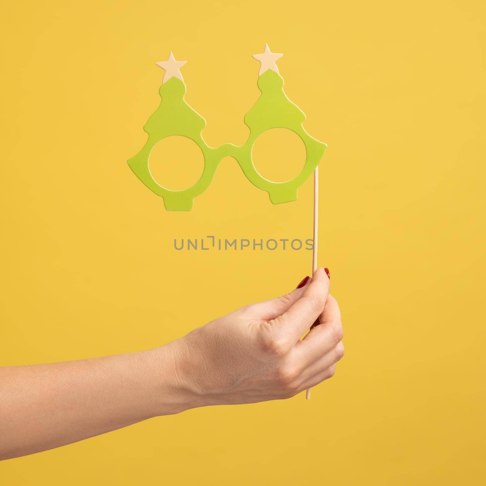 Closeup side view portrait of woman hand holding festive party props, green paper eyeglasses on stick, celebrating holiday. Indoor studio shot isolated on yellow background.