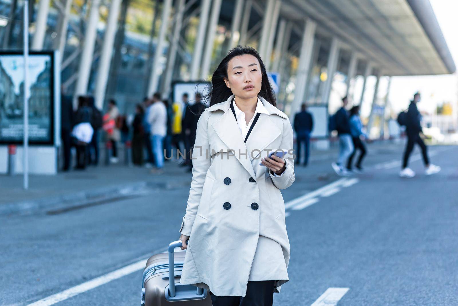 Beautiful female tourist near the airport, a Chinese woman walking with a big suitcase, an Asian woman holding a phone, using the application for booking accommodation and ordering a taxi