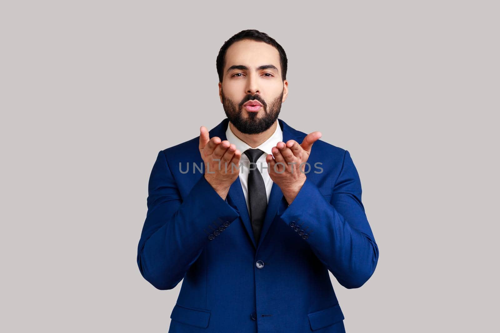 Handsome bearded man sending air kiss over palms, flirting to camera, demonstrating romantic amorous feelings, wearing official style suit. Indoor studio shot isolated on gray background.