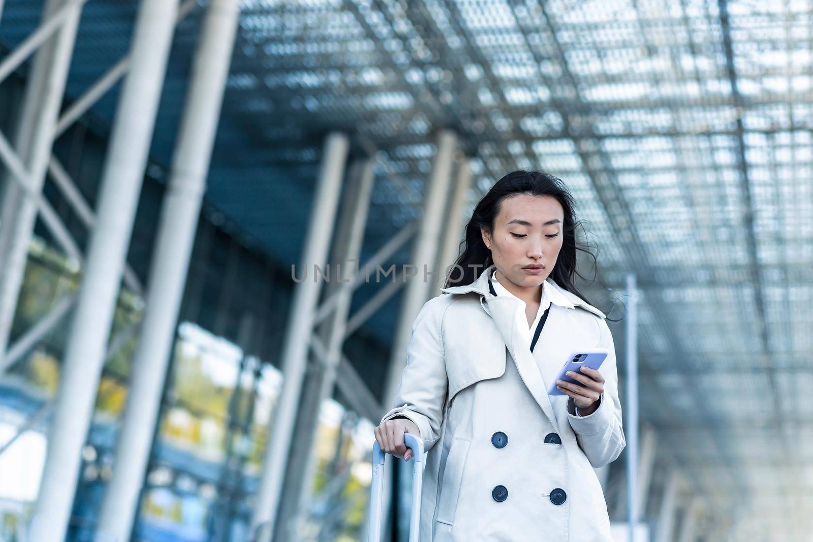 Beautiful female tourist near the airport, a Chinese woman walking with a big suitcase, an Asian woman holding a phone, using the application for booking accommodation and ordering a taxi