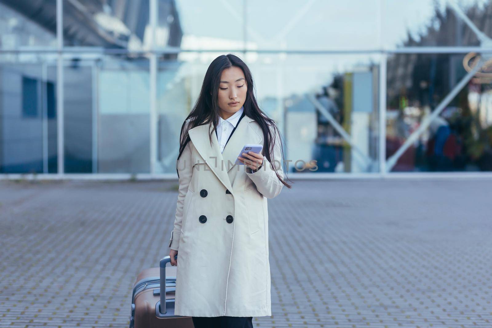 Beautiful female tourist near the airport, a Chinese woman walking with a big suitcase, an Asian woman holding a phone, using the application for booking accommodation and ordering a taxi
