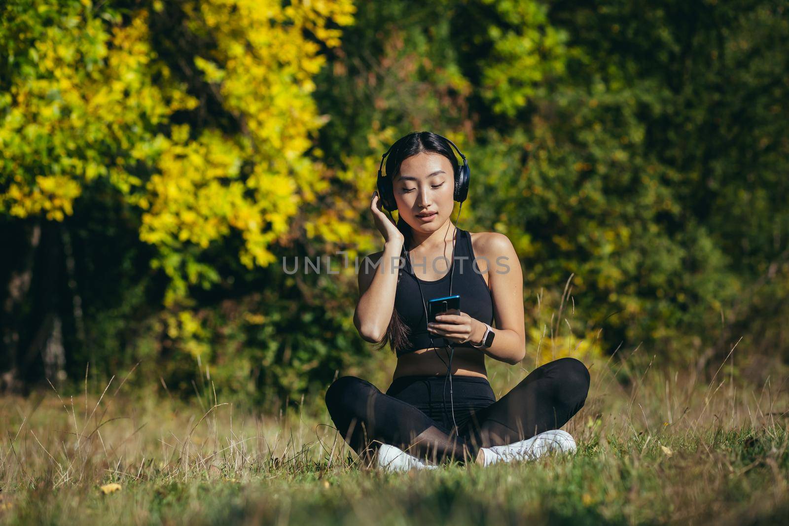 Young beautiful Asian woman sitting in lotus position in the park, relaxing after fitness and running, listening to music on headphones, and using mobile app on phone for fitness