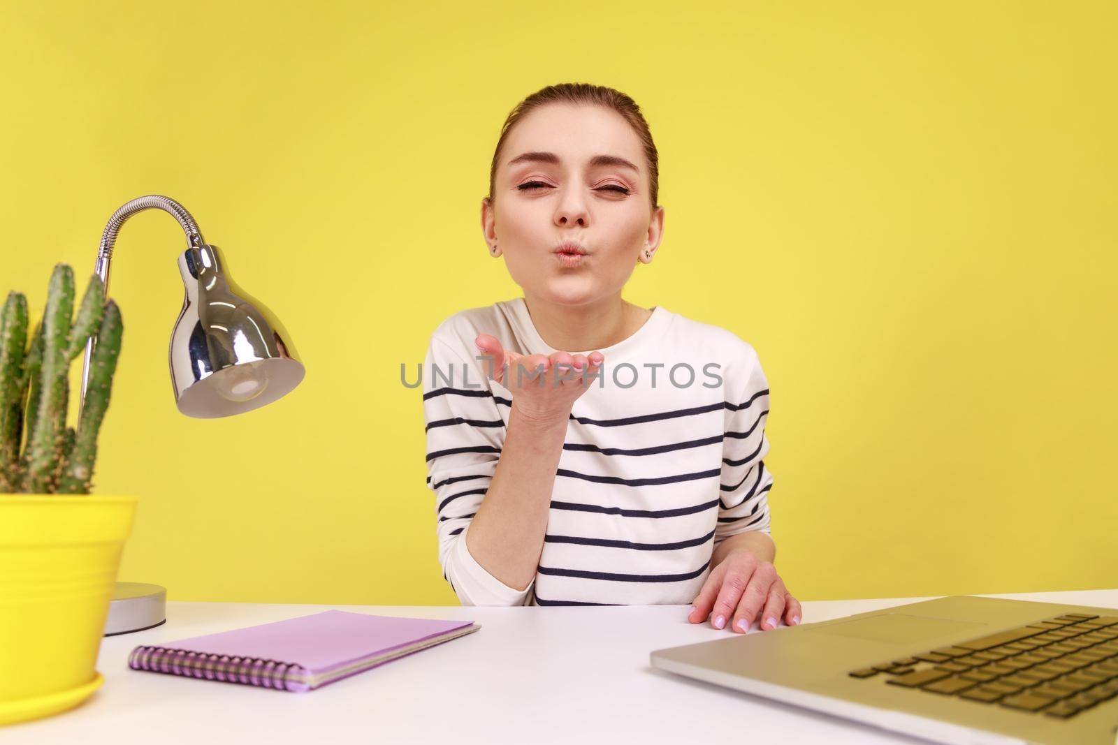 Positive friendly woman making selfie POV, sending air kisses to followers while having livestream, sitting at workplace with laptop. Indoor studio studio shot isolated on yellow background.