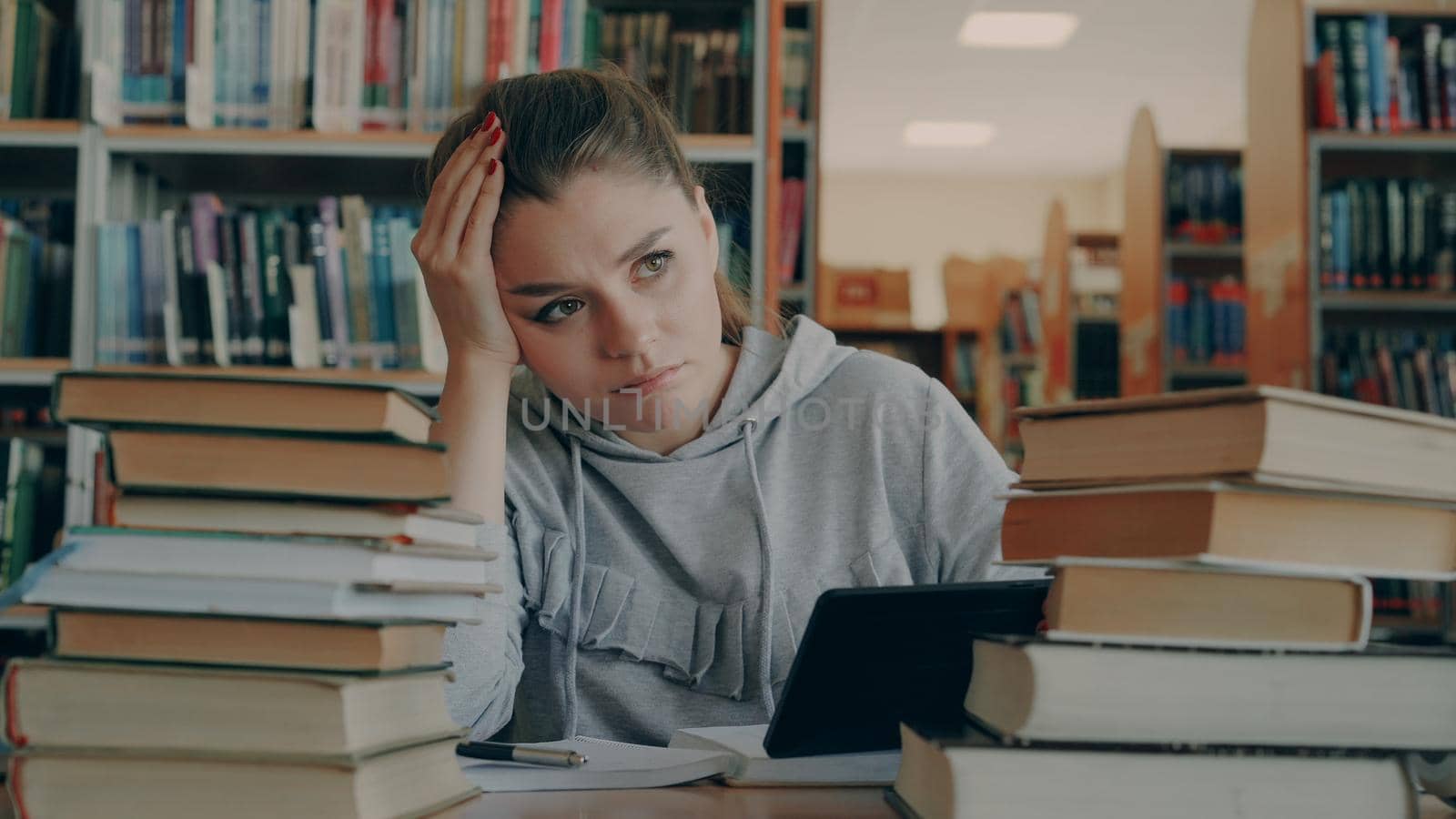 Beautiful caucasian woman is sitting at table in university library surrounded by piles of books holding digital tablet. She is tired and exhausted