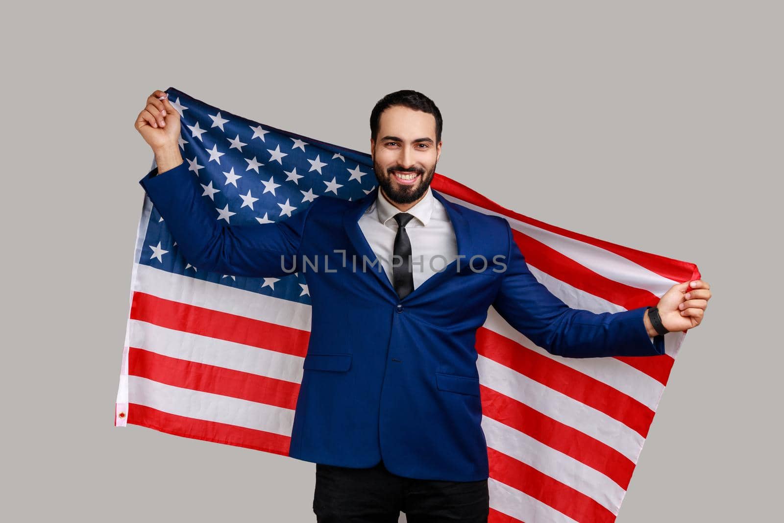 Smiling bearded man holding USA flag and looking at camera with happy look, celebrating national holiday, wearing official style suit. Indoor studio shot isolated on gray background.