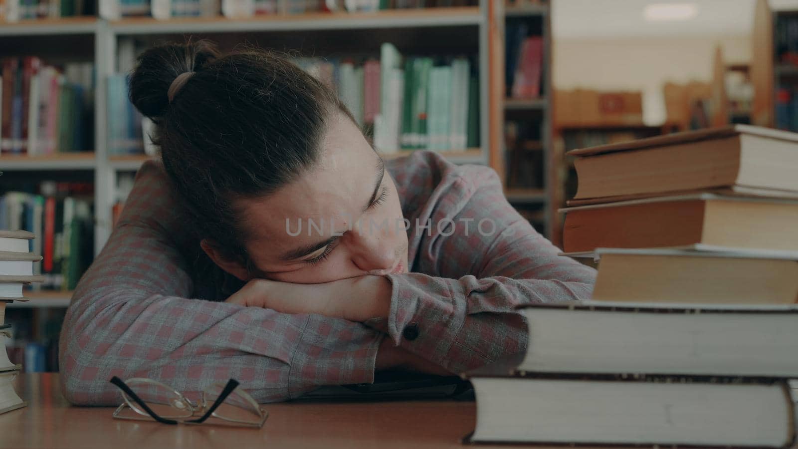 Handsome young caucasian man sitting with his head on table is sleeping in big school library surrounded by huge piles of books. His glasses are lieing in front of him