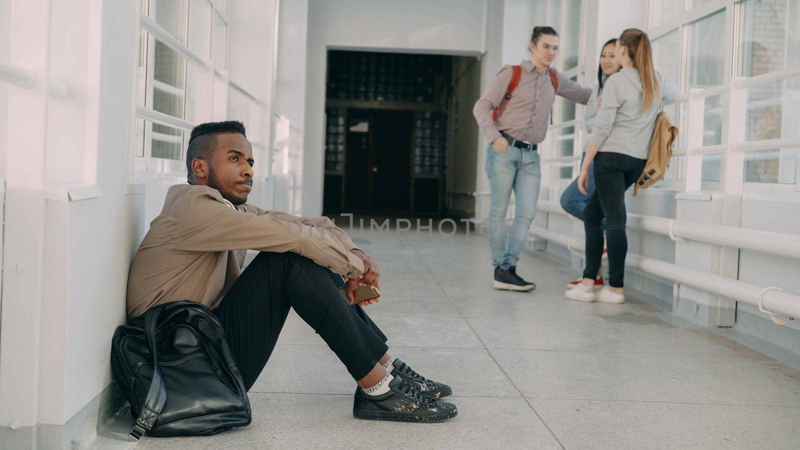 View of upset serious african-american male student sitting on floor in white spacious university corridorwhile his groupmates are standing communicating.