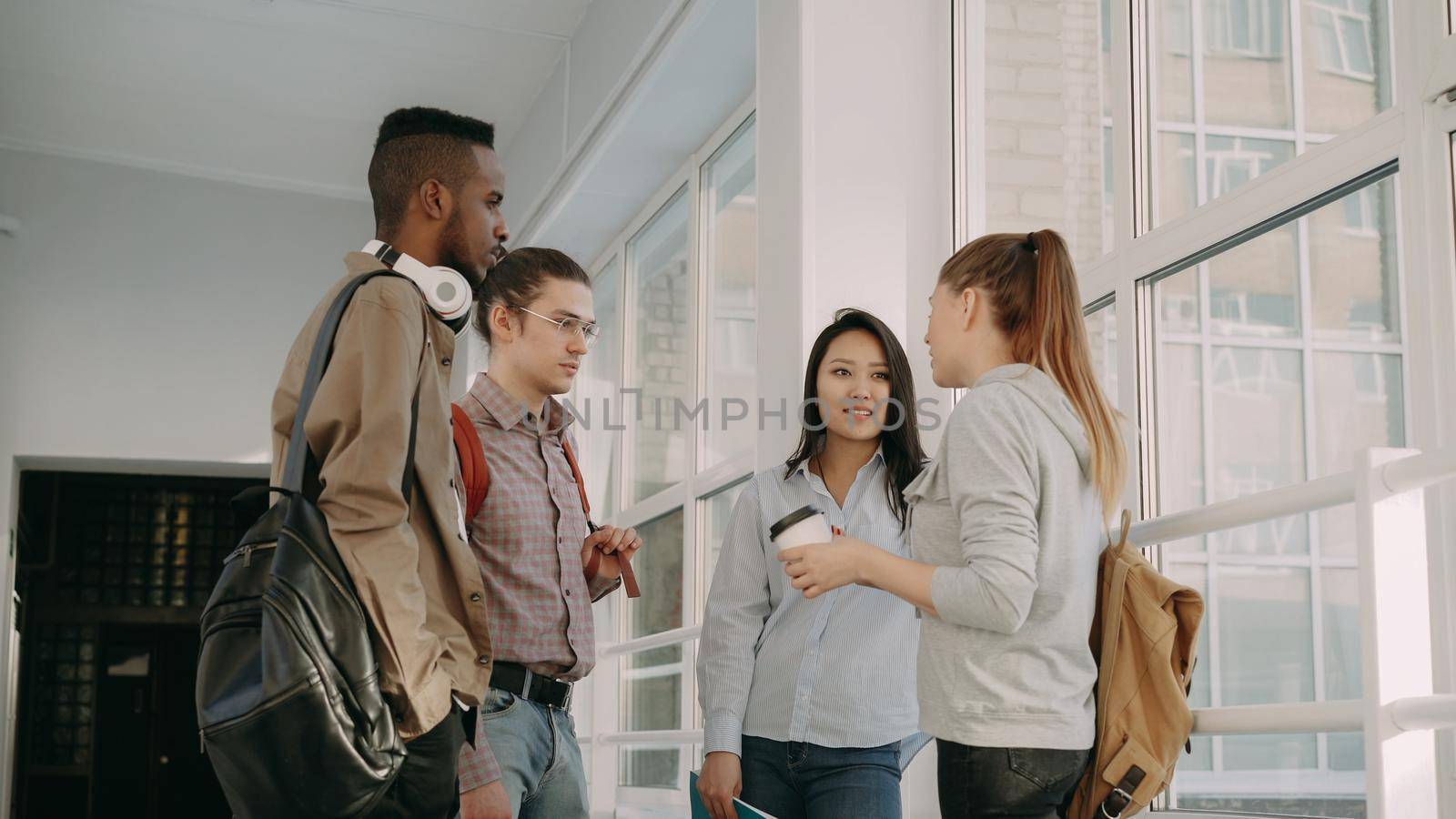 Four multi-ethnic studens are standing in big white spacious hall in college talking to each other in positive way. They are smiling positively and laughing by silverkblack