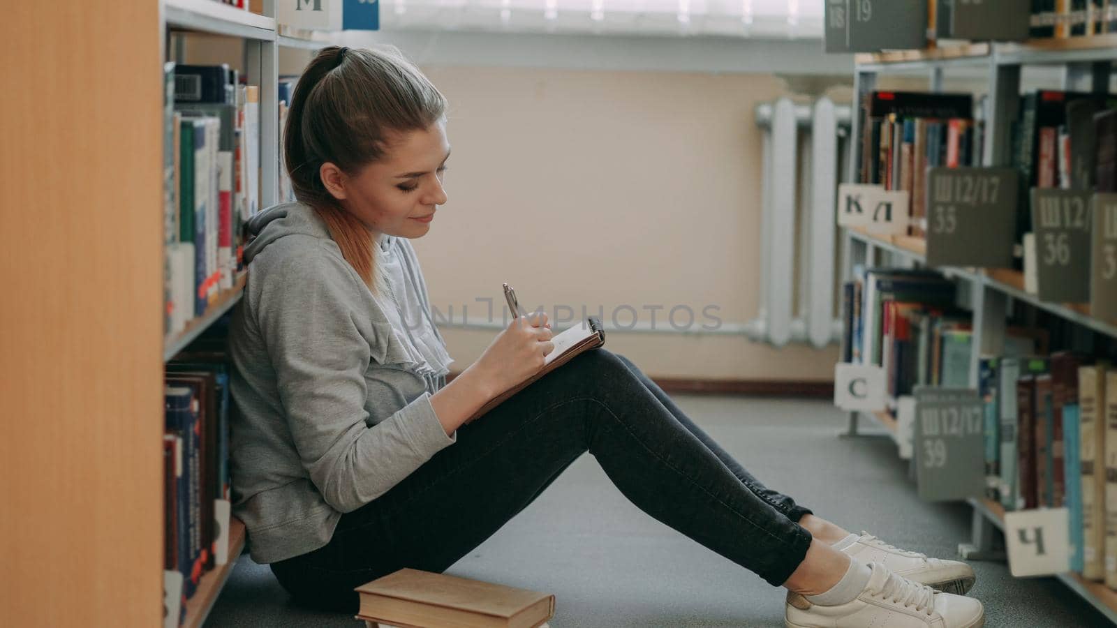Young beautiful dreamy caucasian female student is sitting on floor in big lighty library among bookshelves writing down composition in copybook smiling looking up at ceiling.