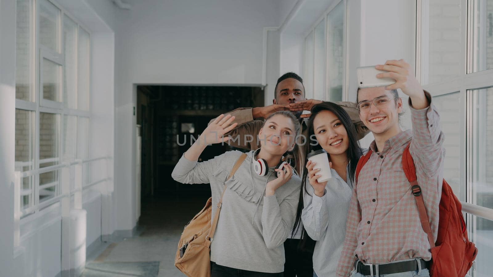 Group of multi-ethnic students taking selfie on smartphone camera while standing in corridor of university . Hipster guy holding phone and friends are posing positively by silverkblack