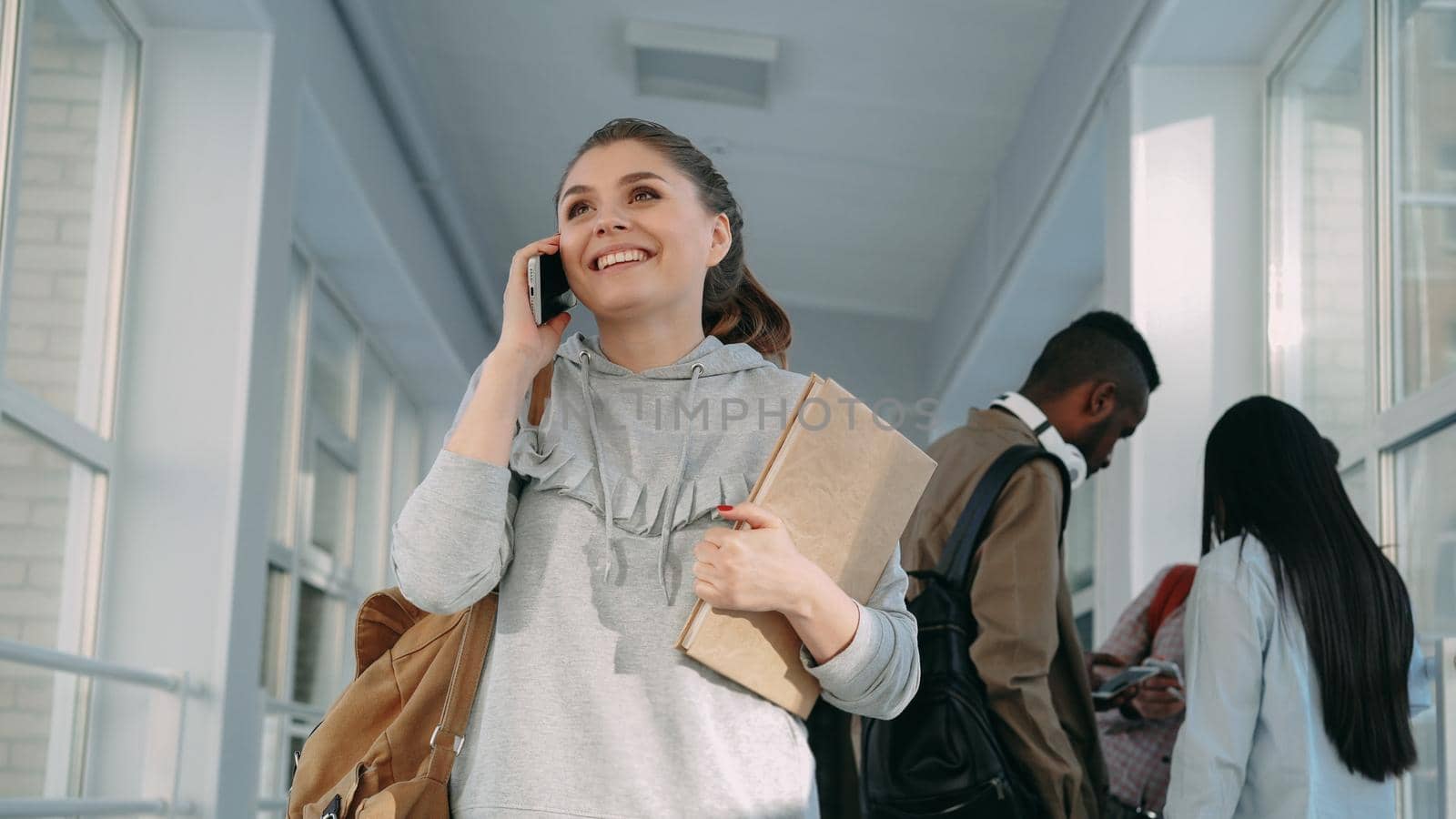 Portrait of young beautiful female student wearing casual clothes talking on phone in positive way while her multi-ethnic groupmates are standing behind her discussing something lively.