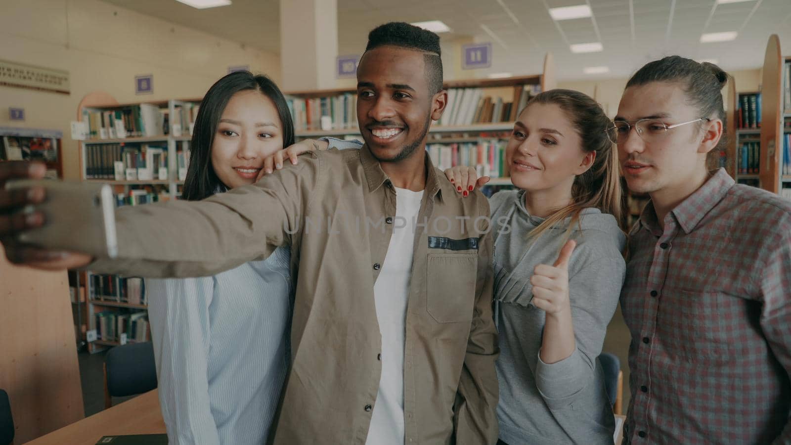 Group of international students have fun smiling and making selfie photos on smartphone camera at university library. Cheerful friends have rest while preapre project together by silverkblack