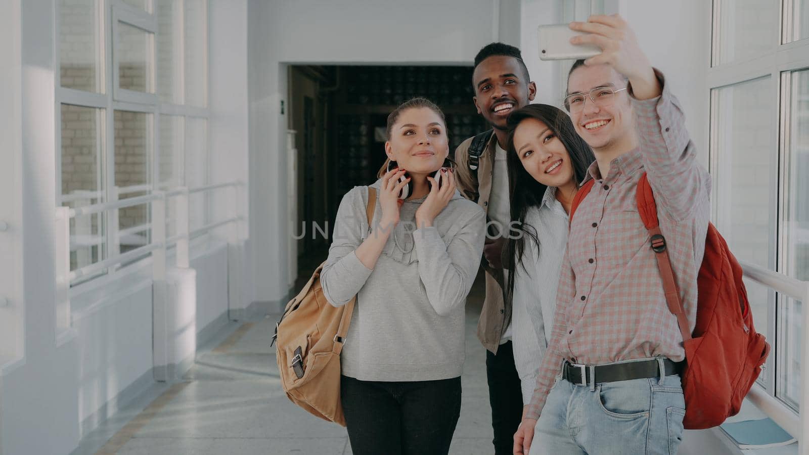 Group of multi-ethnic students taking selfie on smartphone camera while standing in corridor of university . Hipster guy holding phone and friends are posing positively by silverkblack