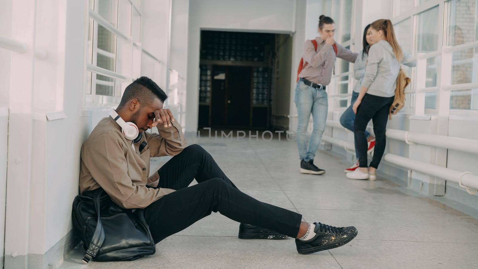 View of upset serious african-american male student sitting on floor in white spacious university corridorwhile his groupmates are standing communicating by silverkblack